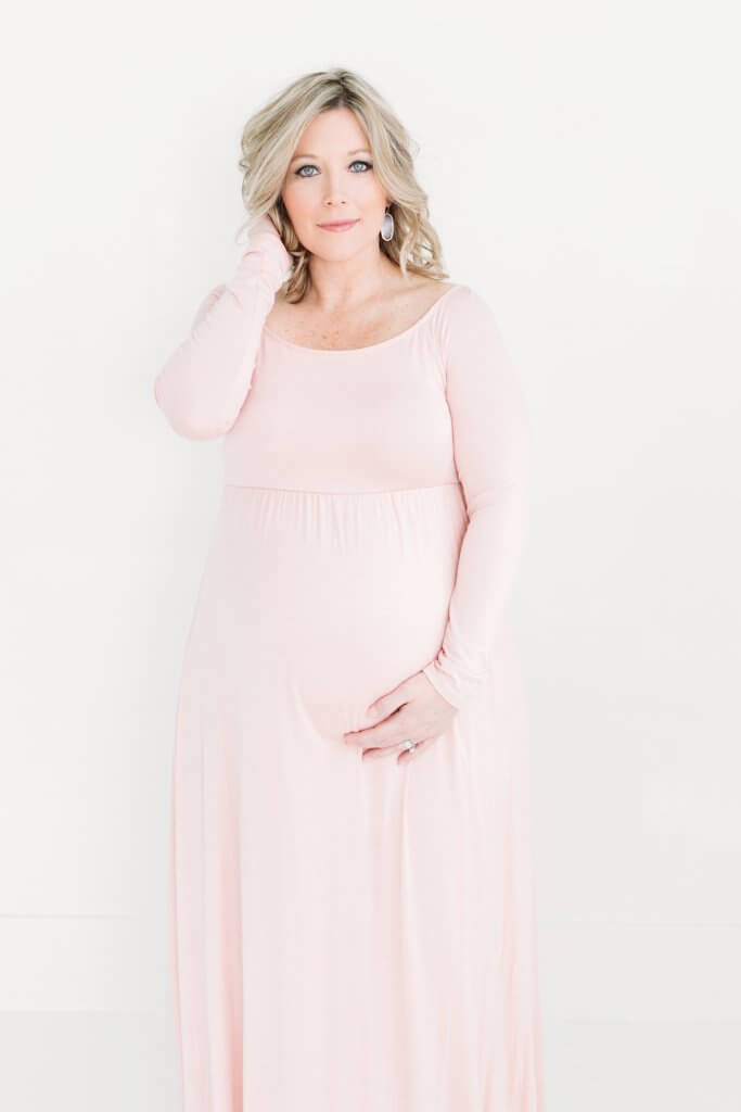 A pregnant woman in a pink dress with her hands in her hair and under her belly