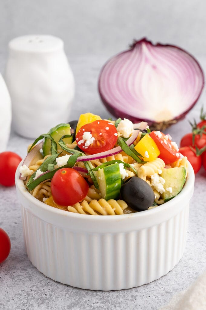 a small white bowl filled with balsamic pasta salad with a red onion that is cut in the background and a salt shaker