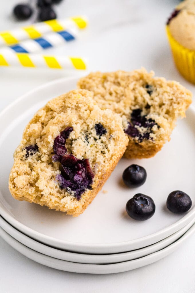 A lemon blueberry muffin on a white plate cut in half with a few blueberries on the plate