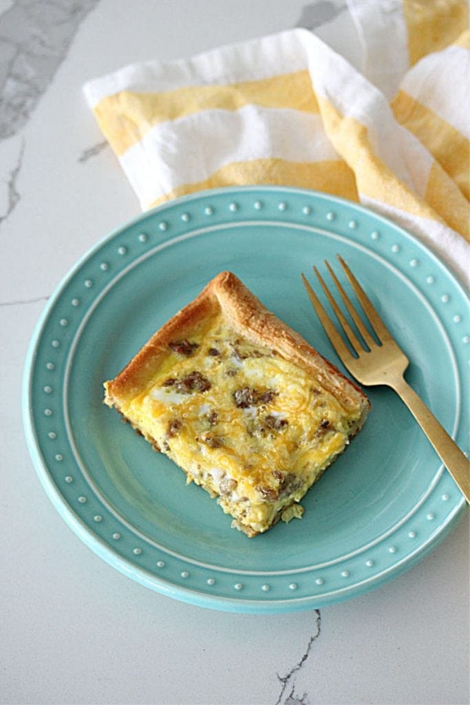 A piece of breakfast casserole on a teal plate with a gold fork and a white and gold napkin
