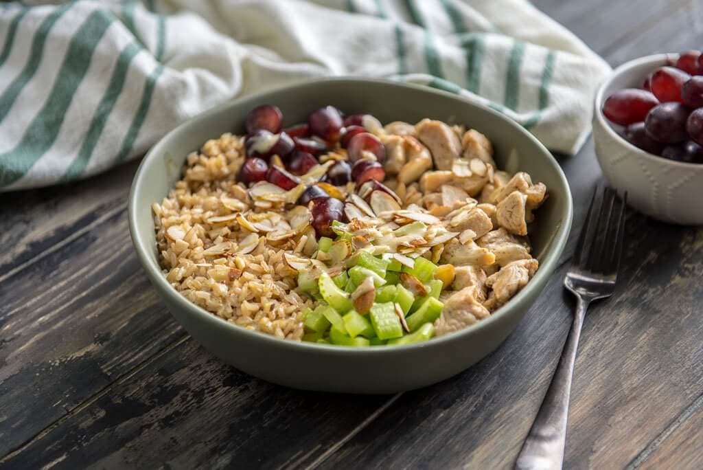 Healthy Chicken Salad Brown Rice Bowl on a wood table with a napkin, fork and bowl of grapes.