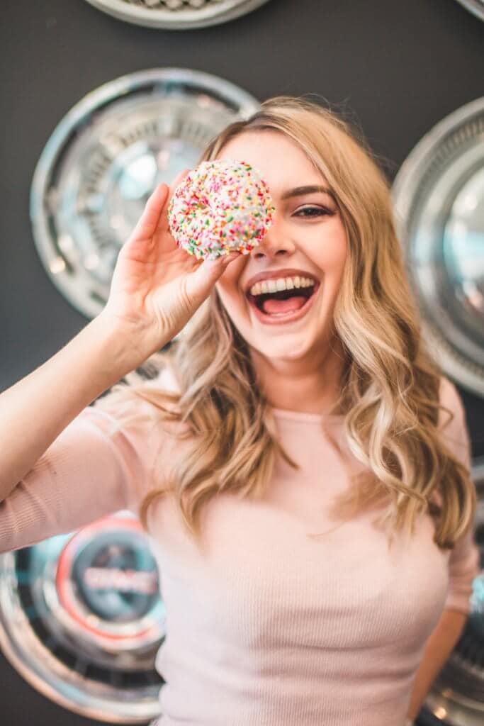 A woman with blonde hair and a pink top holding a donut in front of her face and smiling.