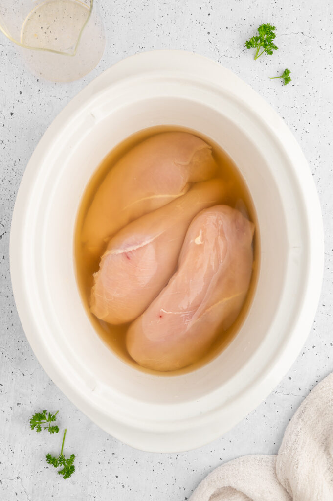 a white slow cooker filled with 3 pieces of raw chicken