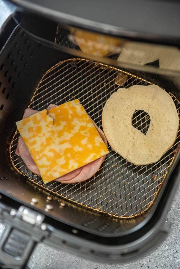Air fryer basket with a bagel in it with ham and cheese on one side of the sliced bagel