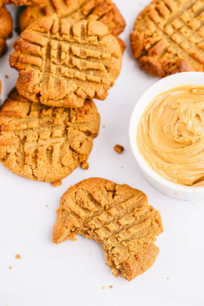a pile of peanut butter cookies, one with a bite taken out of it next to a bowl of peanut butter