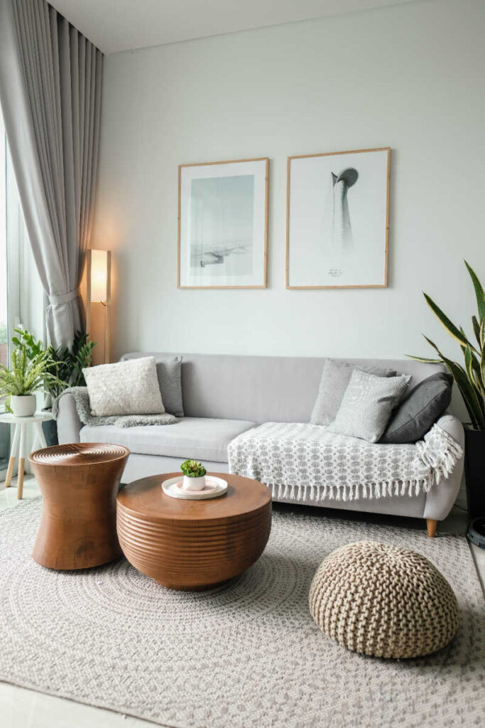 a picture of a living room with plants and a gray couch with white pillows and two wooden coffee tables and a fabric poof