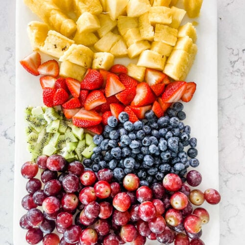 pineapple, strawberries, kiwi, blueberries and grapes on a white cutting board on a marble countertop