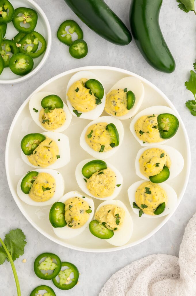 a white plate filled with deviled eggs with jalapenos on top of each one and a few fresh jalapenos and diced jalapenos in a bowl