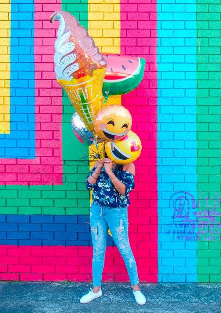 A bright colored wall with a woman holding balloons in front of her face. Smiley face, ice cream and watermelon balloons.