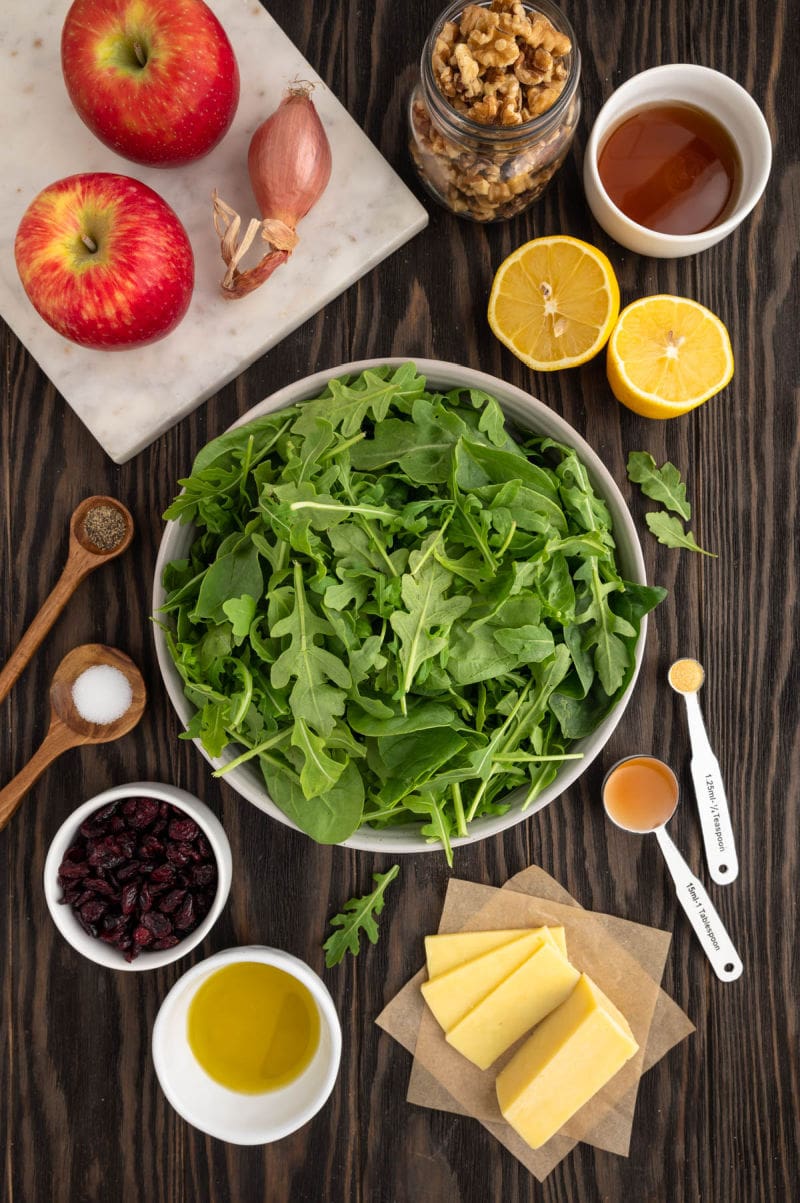 bowl of greens, cranberries, lemon, apple, cheddar, olive oil, on a wooden board