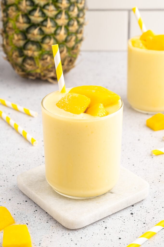 mango pineapple smoothie in a glass cup on a white coaster with a pineapple in the background and a straw in the cup