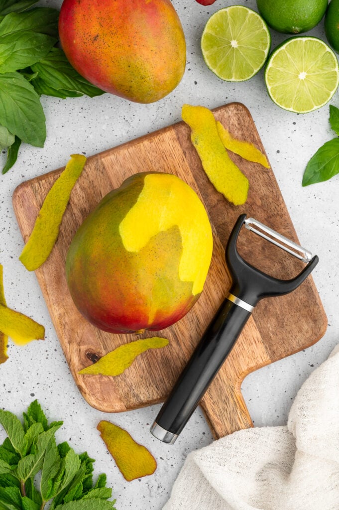 a mango with part of the skin on with a vegetable peeler on a wooden cutting board