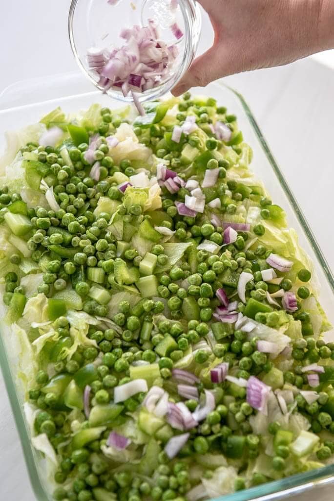 Woman sprinkling onion on top of lettuce and peas in a 9x13 pan
