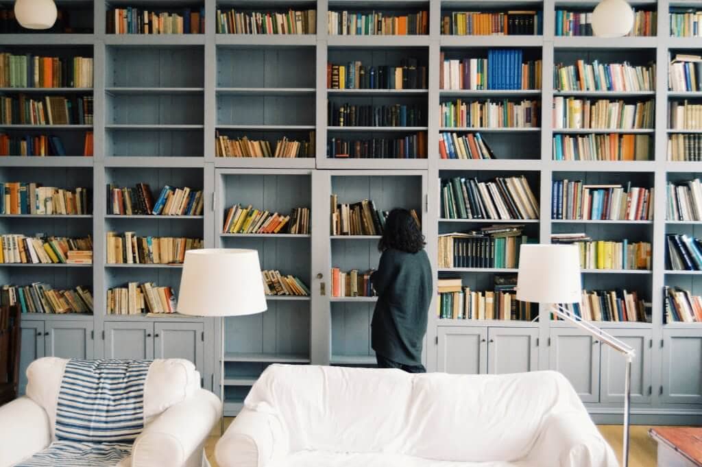 Room full of books on a shelf with a white couch and a woman looking for a book