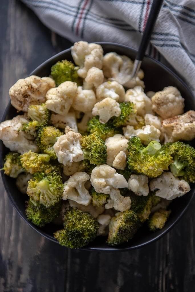 A bowl of roasted broccoli and cauliflower on a wooden board and white napkin