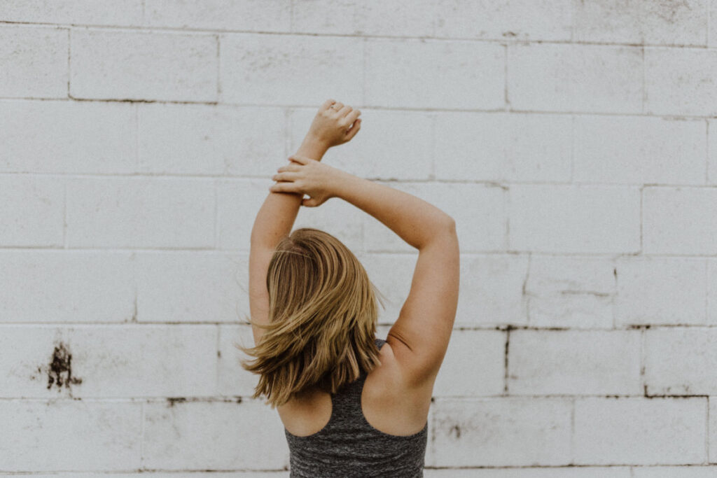 the back of a woman with short blonde hair it a tank top looking at a white brick wall with her hands int he air celebrating