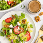 overhead picture of strawberry goat cheese salad with the salad on a plate next to a bowl filled with it and a container of the creamy balsamic dressing next to it