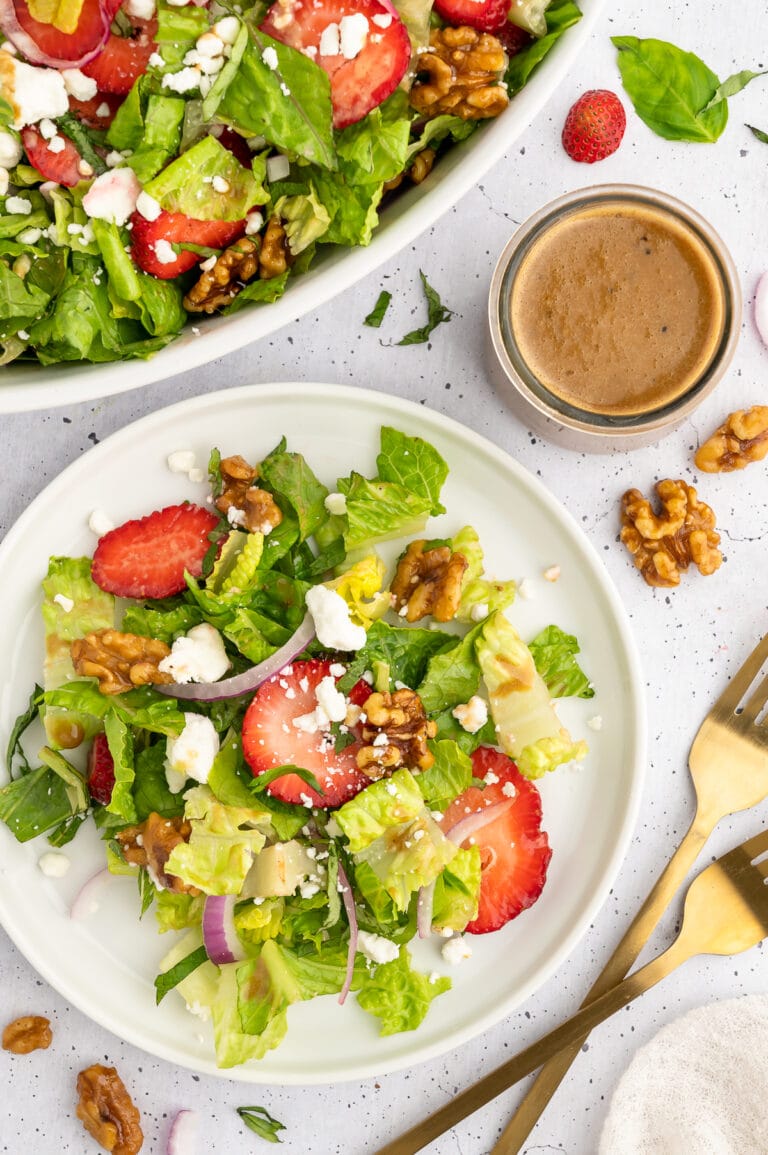 overhead picture of strawberry goat cheese salad with the salad on a plate next to a bowl filled with it and a container of the creamy balsamic dressing next to it