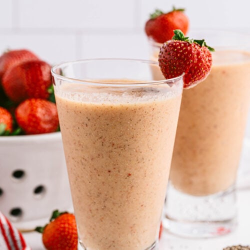 close up photo of two cups of strawberry mango smoothie with strawberries behind them