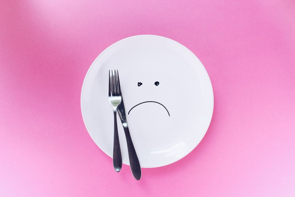 Pink background with a white plate with a sad face painted on it and a knife and fork