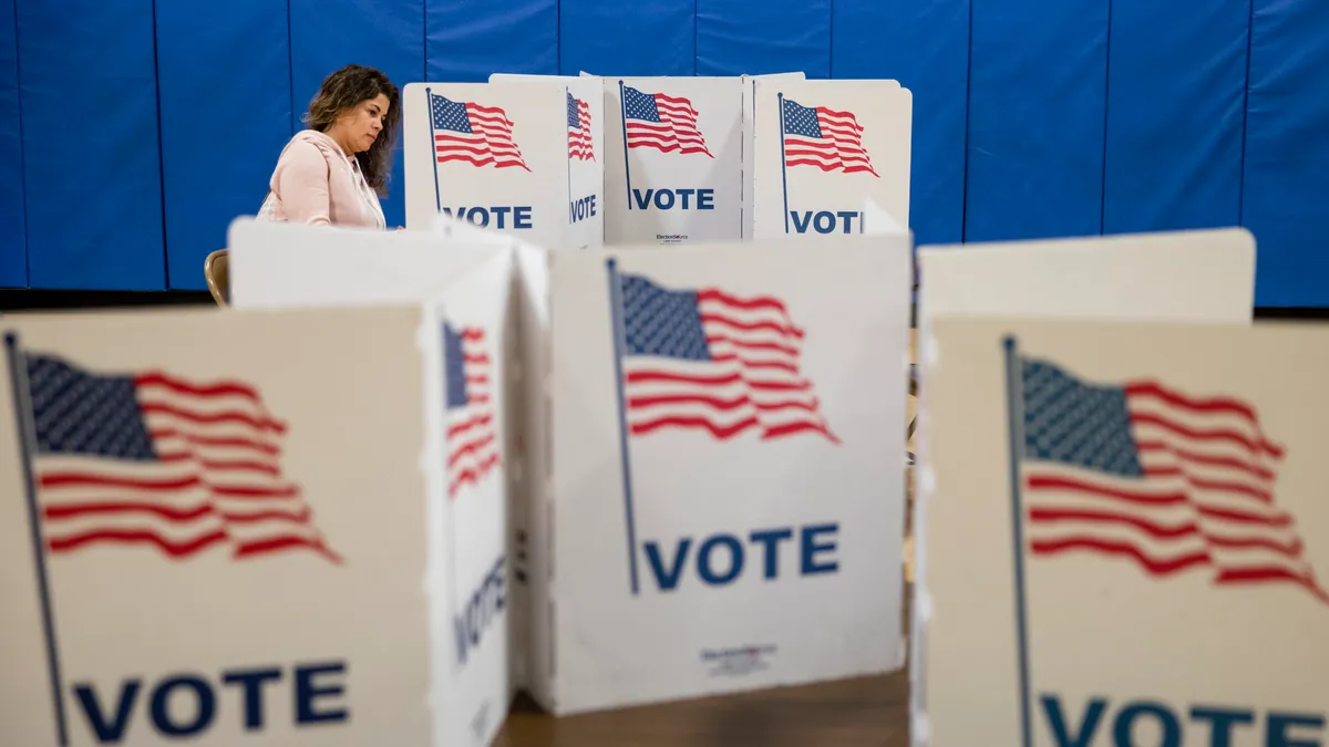 Several voting booths painted white with the American flag and the word VOTE are shown, with a woman in the background casting her vote.