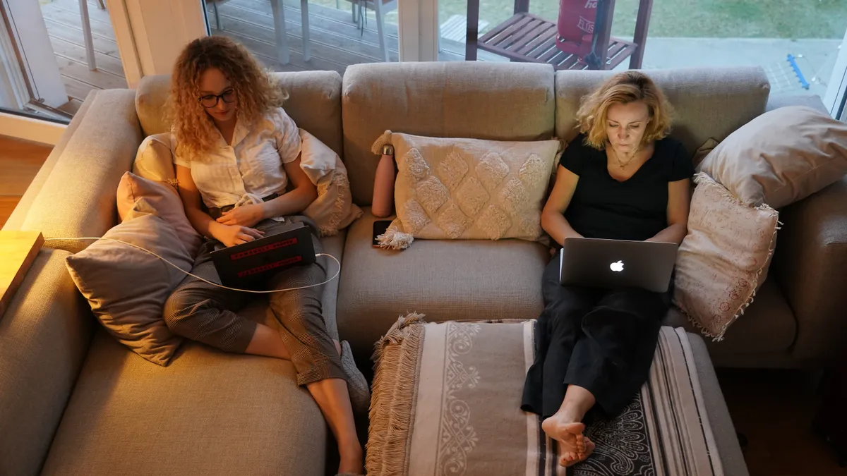 A woman and her daughter sit together on a couch while both scrolling on separate laptops.