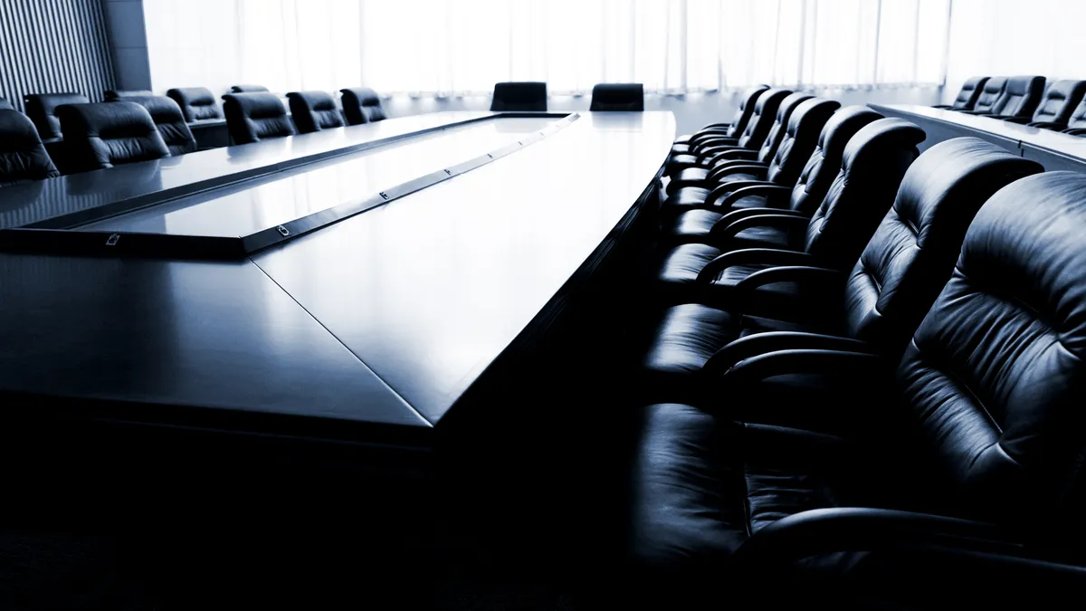 An empty, dark board room is pictured, with chairs surrounding a table.