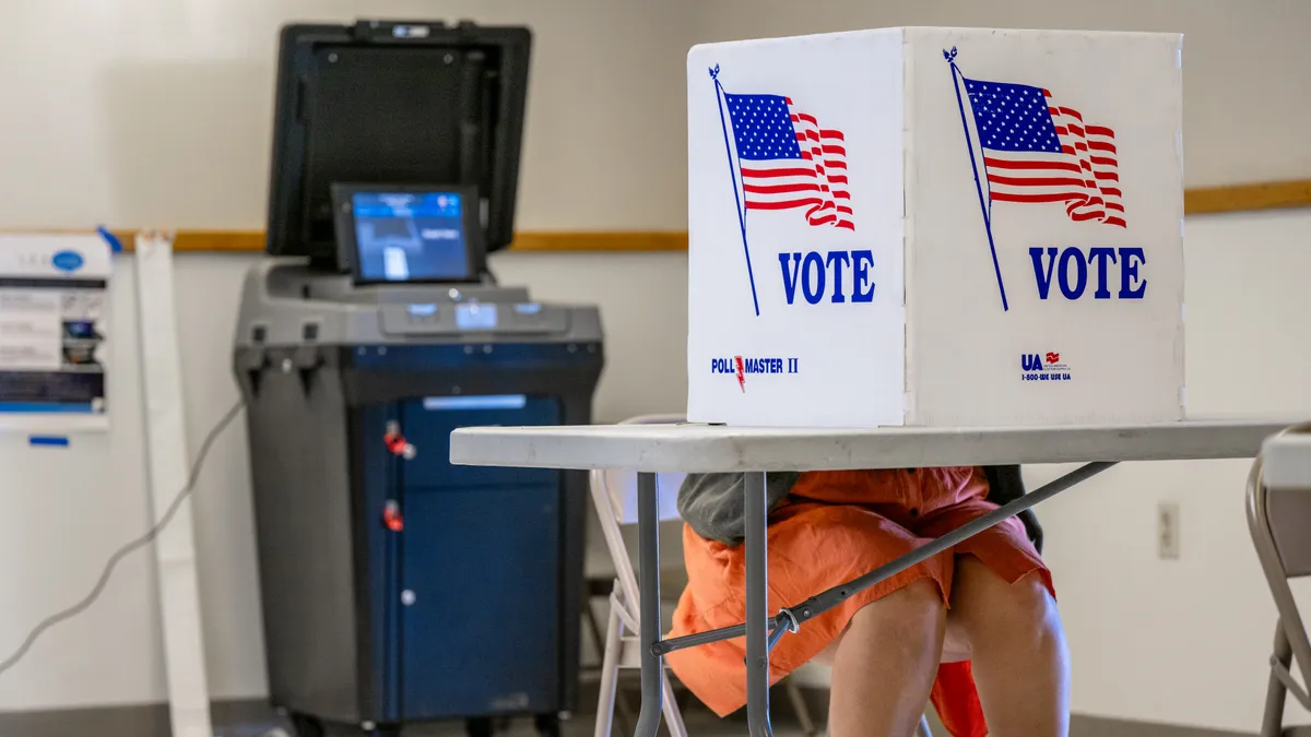 A person sits behind a privacy shield bearing the U.S. flag and the word "vote."