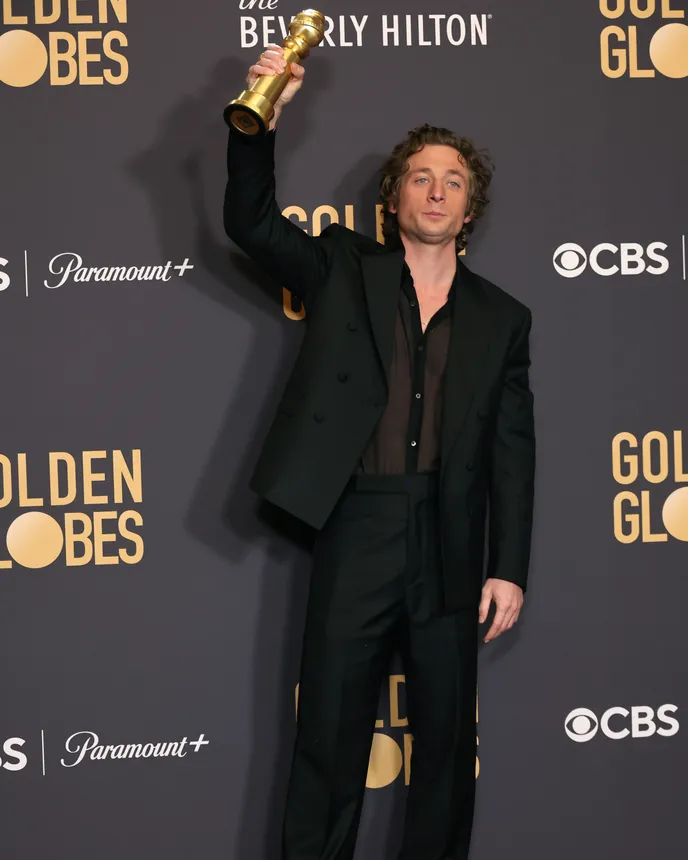 Actor Jeremy Allen White stands on a red carpet, holding his Golden Globe award above his head.