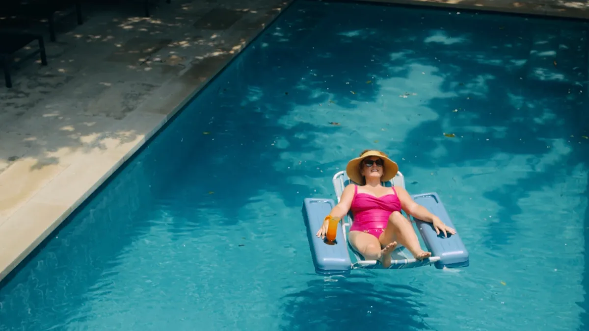 A women in a pink bathing suit and sunhat lays in a blue pool float in the middle of a pool with a cocktail.